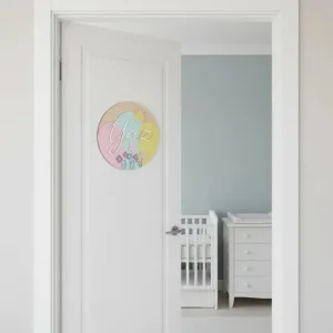 Bahar temalı kapı süsü, mavi duvarlı bebek odasına açılan beyaz kapıda; pastel renk blokları ve örnek isimli tasarım