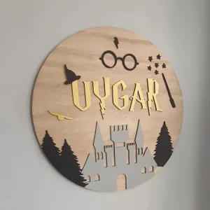 Harry Potter kapı süsünün yandan yakın planı; altın sarısı örnek isim, büyücü şapkası, yıldızlar, gri şato ve çam ağaçları