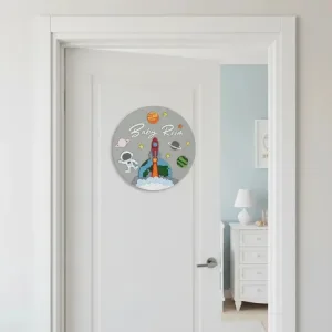 İsimli uzay temalı kapı süsü beyaz kapıda; gri zeminde Baby Room yazısı, roket, astronot, gezegen ve yıldız figürleri