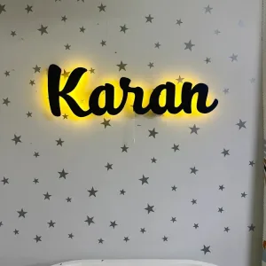 İsme özel lambanın yakın planı: siyah el yazısı harfler, arkadan yayılan sarı ışık ve alttaki açma ipi