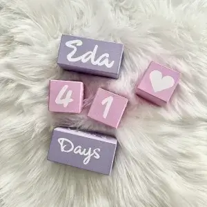 Lila-Pembe-Lila anı küpü seti beyaz tüylü örtüde; beyaz yazılı lila isim ve tarih bloğu, pembe rakam ve kalp küpleri